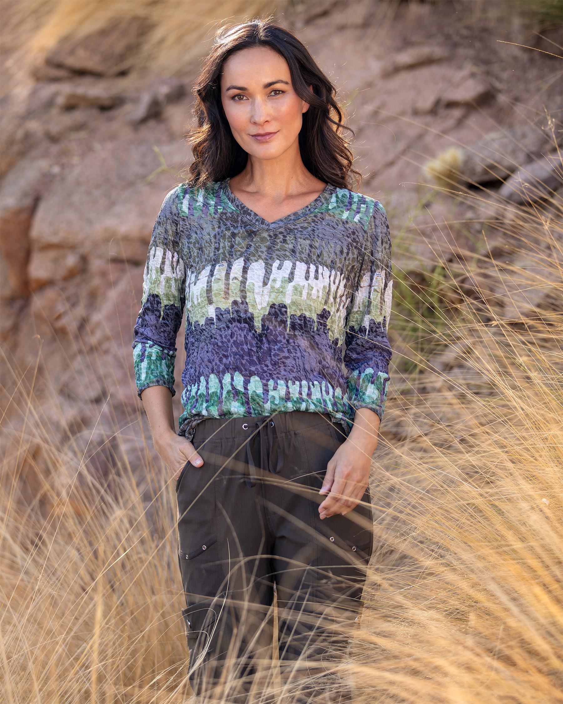 CARINE Fall 2026 lifestyle campaign model wearing a printed top among sunlit grasses and rock.